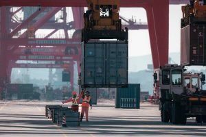 Two workers in orange safety vests loading containers onto a cargo ship.